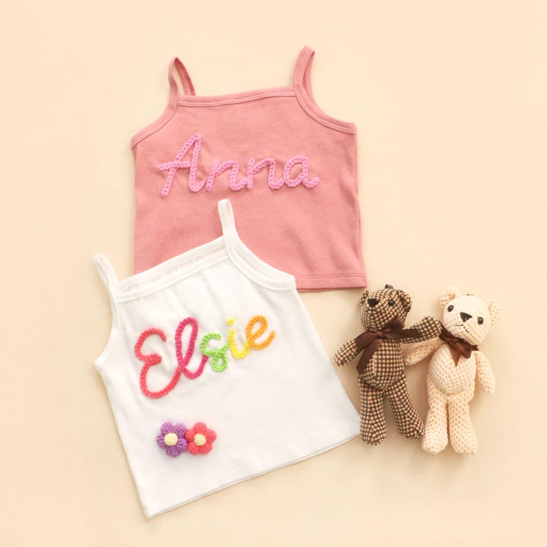May include: Two children's vests, one pink with "Anna" in pink yarn, the other white with "Elsie" in rainbow yarn and flower accents. Two small teddy bears with brown bows are also in the image.