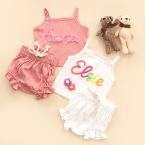 May include: Children's clothing sets featuring pink and white tank tops and shorts. The pink top is embroidered with "Anna", the white one with "Elsie" in rainbow thread. Two small teddy bears are also included. The shorts have a frilled hem and drawstring waist.