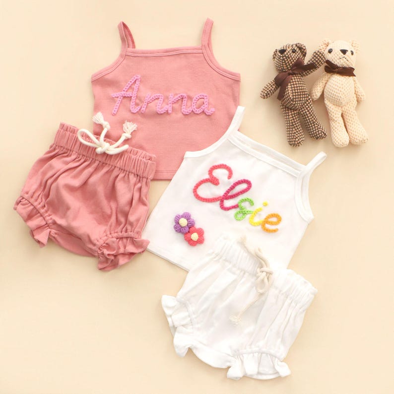 May include: Children's clothing sets featuring pink and white tank tops and shorts. The pink top is embroidered with "Anna", the white one with "Elsie" in rainbow thread. Two small teddy bears are also included. The shorts have a frilled hem and drawstring waist.