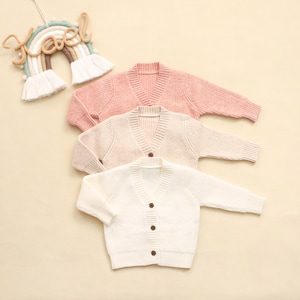 K&ouml;nnte beinhalten: Drei V-Ausschnitt-Cardigans in sanften Pastellfarben: Rosa, Creme und Wei&szlig;. Jeder Pullover hat lange &Auml;rmel und Holzkn&ouml;pfe. Im Hintergrund h&auml;ngt eine regenbogenf&ouml;rmige Wanddekoration mit dem Wort "Noel".