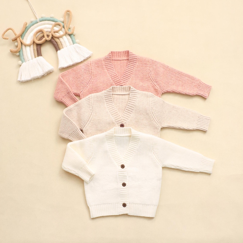 K&ouml;nnte beinhalten: Drei V-Ausschnitt-Cardigans in sanften Pastellfarben: Rosa, Creme und Wei&szlig;. Jeder Pullover hat lange &Auml;rmel und Holzkn&ouml;pfe. Im Hintergrund h&auml;ngt eine regenbogenf&ouml;rmige Wanddekoration mit dem Wort "Noel".