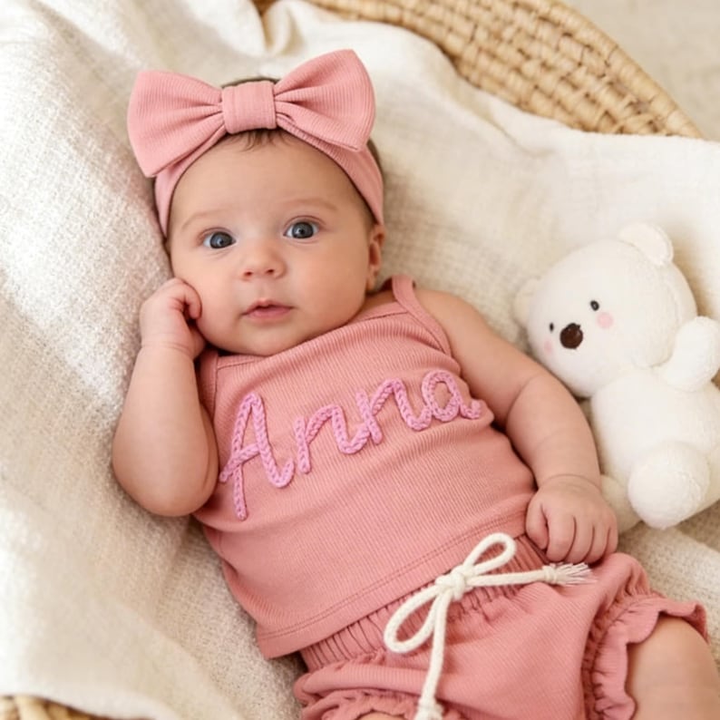May include: A baby in a pink outfit, comprising a tank top embroidered with the name "Anna", matching shorts, and a bow headband. A white teddy bear is beside the baby. The baby is lying on a white blanket in a wicker basket.