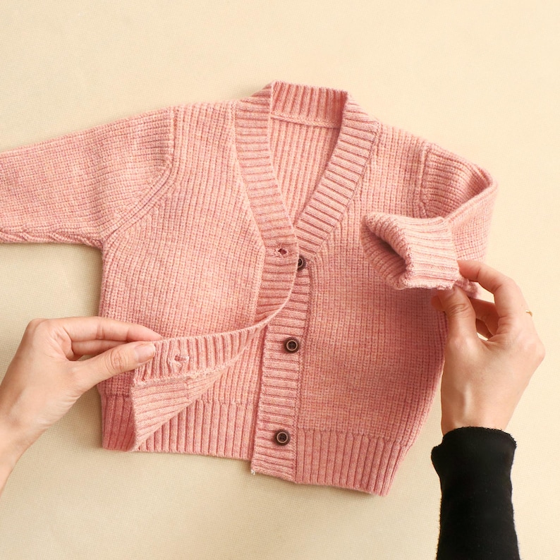 K&ouml;nnte beinhalten: Hellrosa Strickjacke mit Rippenmuster, V-Ausschnitt und drei braunen Kn&ouml;pfen. Der lang&auml;rmlige Pullover wird auf einer hellbeigen Oberfl&auml;che pr&auml;sentiert. Die Strickjacke wird von zwei H&auml;nden offen gehalten.