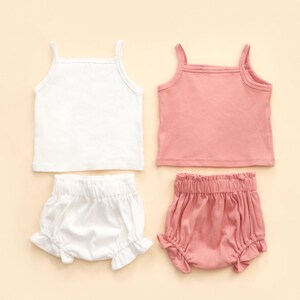 May include: Two sets of children's clothing, each comprising a vest top and ruffled bloomers. One set is white, the other is a soft pink. The vest tops have thin straps and a square neckline. The bloomers have an elasticated waistband.