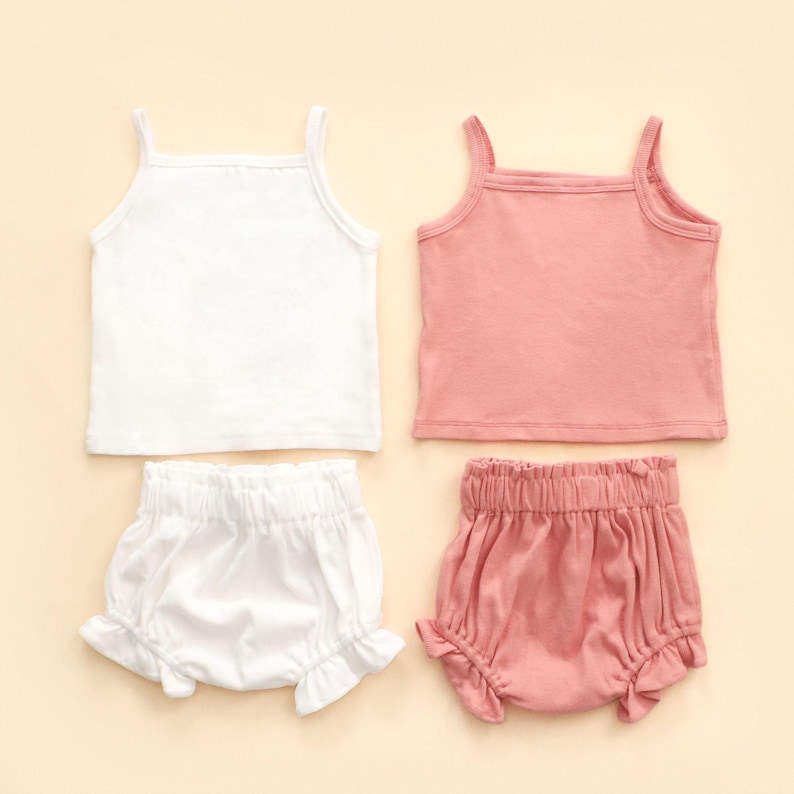 May include: Two sets of children's clothing, each comprising a vest top and ruffled bloomers. One set is white, the other is a soft pink. The vest tops have thin straps and a square neckline. The bloomers have an elasticated waistband.