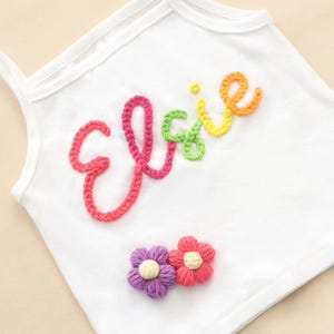 May include: White vest top with the name "Elsie" embroidered in colourful yarn. The name is in a rainbow of colours, with pink, green, yellow, and orange. Two small crocheted flowers in purple and red are below the name.