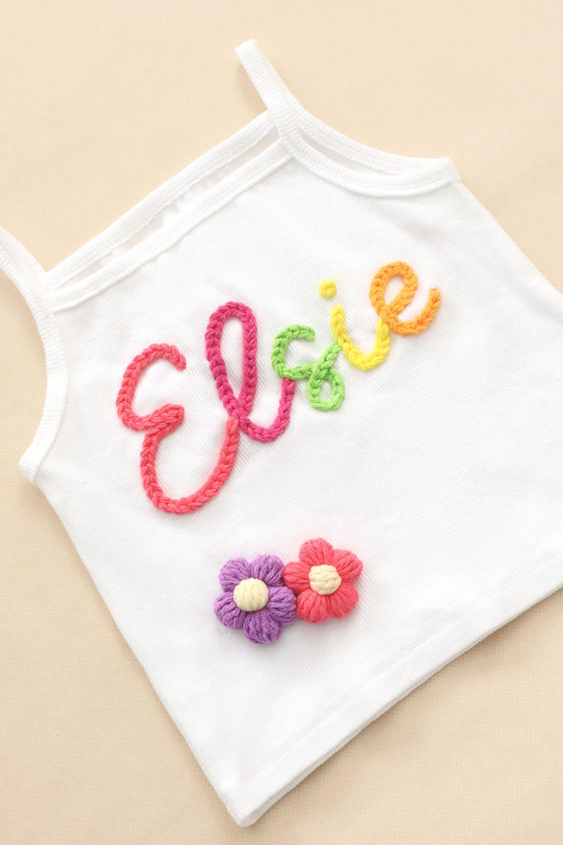 May include: White vest top with the name "Elsie" embroidered in colourful yarn. The name is in a rainbow of colours, with pink, green, yellow, and orange. Two small crocheted flowers in purple and red are below the name.