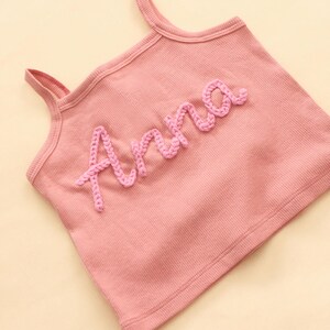 May include: A pink ribbed vest top with shoulder straps. The name "Anna" is embroidered in a lighter pink, cursive font across the front. The top is made of a soft, comfortable material.