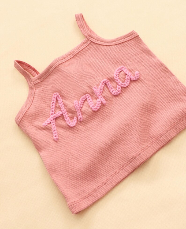 May include: A pink ribbed vest top with shoulder straps. The name "Anna" is embroidered in a lighter pink, cursive font across the front. The top is made of a soft, comfortable material.