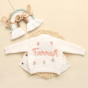 K&ouml;nnte beinhalten: Cremefarbene Baby-Strickjacke mit dem Namen "Farrah" in Rosa gestickt, verziert mit kleinen rosa Blumenakzenten. Im Hintergrund ist ein regenbogenf&ouml;rmiger Wandbehang mit dem Namen "Noel" zu sehen.