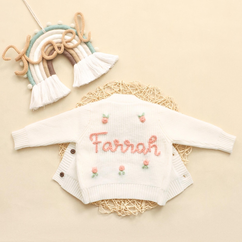 K&ouml;nnte beinhalten: Cremefarbene Baby-Strickjacke mit dem Namen "Farrah" in Rosa gestickt, verziert mit kleinen rosa Blumenakzenten. Im Hintergrund ist ein regenbogenf&ouml;rmiger Wandbehang mit dem Namen "Noel" zu sehen.