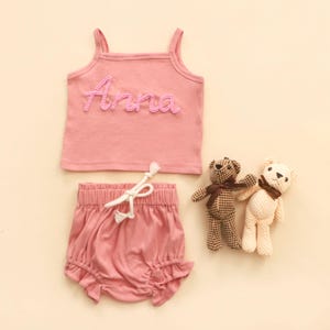 May include: A pink baby clothing set. It includes a tank top with the name "Anna" in pink lettering, matching ruffled shorts, and two small teddy bears. The set is made of soft material.