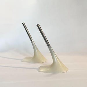 Pode incluir: Duas esculturas abstratas com um design minimalista. Cada escultura apresenta uma base creme que se curva para cima, sustentando uma haste de metal prateado. Os objetos estão sobre um fundo branco liso.