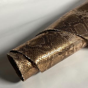 Puede incluir: Hoja enrollada de cuero de color bronce metálico. El cuero tiene un patrón texturizado en forma de escamas. Los bordes del cuero enrollado son visibles, revelando las capas. El fondo es una superficie blanca lisa.