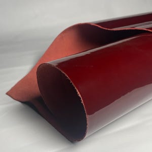 May include: A rolled sheet of burgundy leather with a glossy finish. The underside is a matte, reddish-brown. The material is likely for crafting or upholstery. The leather is a deep, rich colour.