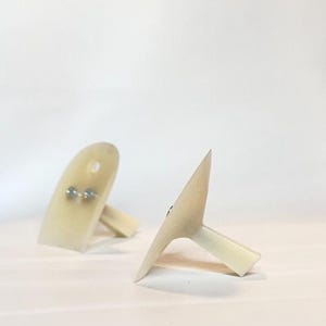 Pode incluir: Dois objetos de plástico esbranquiçados em forma de unha com pequenos fixadores de metal. Cada um tem uma base plana e um topo pontiagudo e curvo. Os objetos estão sobre um fundo branco liso.