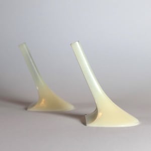 Op de afbeelding: Twee crèmekleurige, abstract gevormde objecten met een gebogen ontwerp. De objecten hebben een brede basis die taps toeloopt in een dunne, langwerpige top. Ze staan tegen een effen, lichte achtergrond.