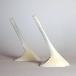 May include: Two white, abstract shoe heels with a curved design. The heels are angled and appear to be made of a smooth, glossy material. The heels are set against a plain, light background.