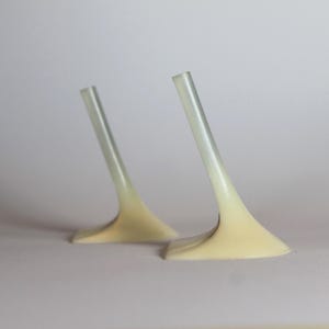 Op de afbeelding: Twee abstracte, minimalistische sculpturen met een lichte, doorschijnende steel en een uitlopende, gebroken witte basis. De stelen zijn schuin geplaatst, wat een gevoel van beweging creëert. De achtergrond is zachtgrijs.