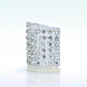 May include: A clear, rectangular shoe heel with a grid pattern of sparkling, round, clear rhinestones. The heel has a slight angle at the top and sits on a clear base, reflecting light. The overall design is elegant and glamorous.
