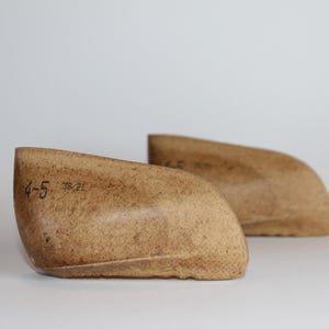 May include: Two light brown shoe lasts, side by side, against a white background. The lasts are made of a textured material and have the numbers "4-5" and "38/21" printed on them. The shoe lasts are used for shaping shoes.