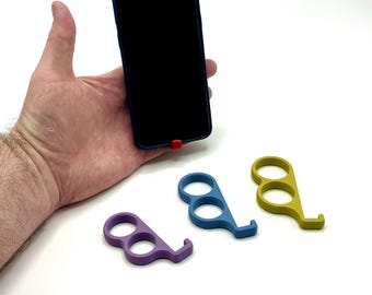 Puede incluir: Un teléfono inteligente en una funda azul con un acento rojo se sostiene en una mano. Se muestran tres herramientas multicolores en forma de gancho, del tamaño de un dedo: morado, azul y amarillo. Las herramientas están diseñadas para evitar tocar superficies.
