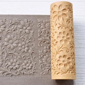Pode incluir: Um rolo de madeira com um padrão floral ao lado de uma placa de argila cinza com o mesmo design floral em relevo. O rolo é cilíndrico e apresenta um padrão repetido de flores e folhas. A placa de argila mostra a impressão do design do rolo.