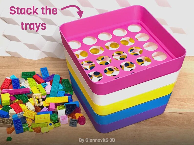 May include: A stackable toy storage system with a pink top tray and multiple colored trays below. The top tray has circular cutouts. The text "Stack the trays" is visible. A pile of colorful toy blocks is next to the storage system.