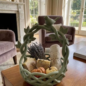 Op de afbeelding: Een decoratieve groene keramische mand met een vogel op het handvat, gevuld met lavendel, zeep, een opgerolde handdoek en kleine pompoenen. De mand staat op een houten tafel.