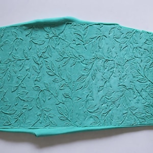 May include: A teal-colored, leaf-patterned rolling pin with matching embossed dough. Two wooden rolling pins with leaf patterns are on either side of the dough. The rolling pin is likely used for baking or crafting.