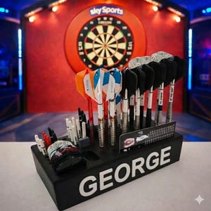 Może przedstawiać: Czarny uchwyt na rzutki z białym napisem "GEORGE", trzymający wiele rzutek z lotkami w różnych kolorach. W tle tarcza do darta. Rzutki mają srebrne korpusy i kolorowe lotki.