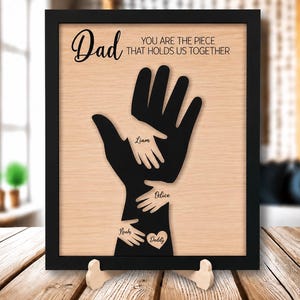 Puede incluir: Un letrero de madera enmarcado con la palabra "Dad" en cursiva y la frase "You are the piece that holds us together". Una silueta de mano negra sostiene recortes de manos más pequeños con nombres. Un corazón con "Daddy" está en la parte inferior.