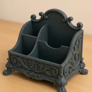 May include: A dark teal desk tidy with four compartments and elaborate scrollwork. The organiser has a curved back and sits on small, decorative feet. The design is reminiscent of antique furniture.