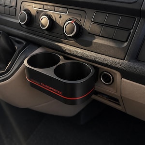 May include: Black dual cup holder with a red trim, installed in a vehicle's dashboard. The cup holder is labelled "TRANSPORTER" in red. The dashboard includes controls, vents, and a USB port.