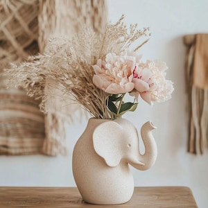 May include: A beige ceramic elephant-shaped vase filled with pink peonies and dried grasses. The vase features an elephant head design with a trunk and ear. The flowers provide a splash of colour against the neutral background.