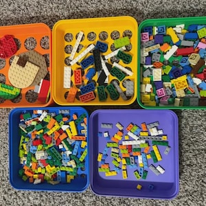 May include: An overhead shot of five colorful plastic bins filled with various toy building blocks. The bins are orange, yellow, green, blue, and purple. The blocks are in various colors and sizes, including red, white, blue, and yellow.
