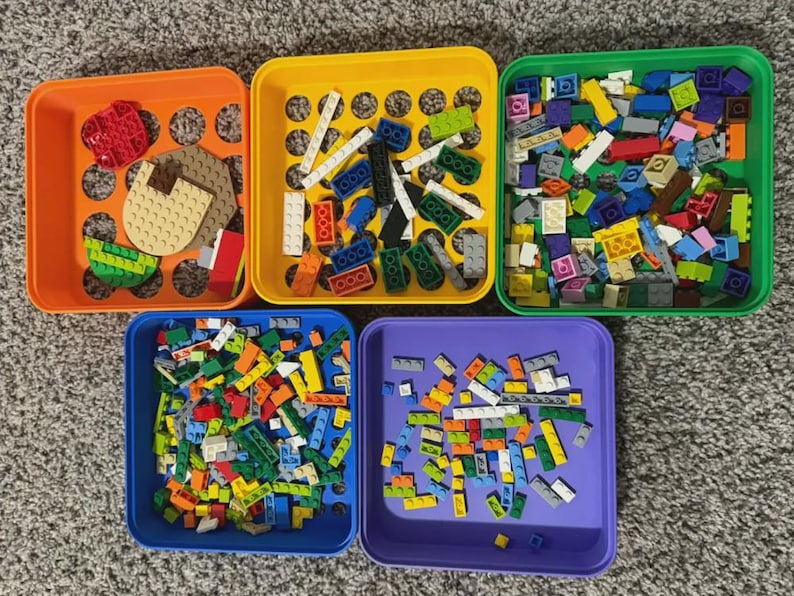 May include: An overhead shot of five colorful plastic bins filled with various toy building blocks. The bins are orange, yellow, green, blue, and purple. The blocks are in various colors and sizes, including red, white, blue, and yellow.