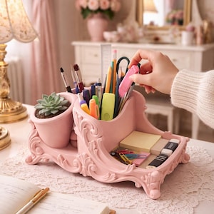 Pode incluir: Um organizador de mesa rosa com uma planta suculenta, porta-canetas e uma pequena bandeja para materiais. O organizador tem um design ornamentado e vintage e é preenchido com canetas, lápis e notas adesivas. Uma mão alcança uma borracha rosa.