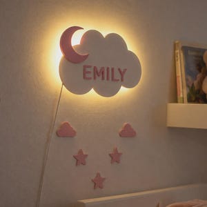 Könnte beinhalten: Eine wolkenförmige Wandleuchte mit dem Namen "EMILY" in rosa Schrift. Ein rosa Halbmond ist an der Wolke befestigt. Zusätzliche rosa Wolken- und Stern-Dekorationen befinden sich darunter. Ein Teddybär und Bücher stehen auf einem Regal.