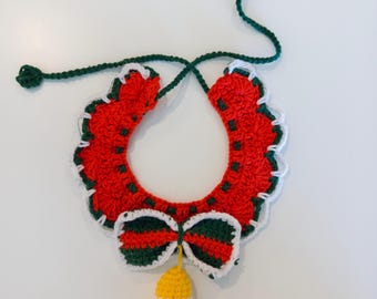 May include: A crochet collar featuring a red scalloped base with dark green accents and a white trim. A bow with alternating red and green stripes is attached at the center, with a small yellow crochet bell hanging from the bottom of the bow.