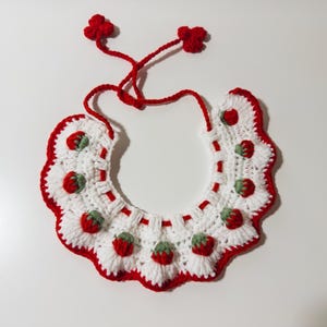 May include: A handmade crochet bib in white with a red scalloped edge. The bib features red and green strawberry appliqués and red ties with small flower accents. A charming accessory.
