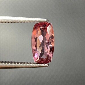 Könnte beinhalten: Ein facettierter, oval geformter Edelstein in Rosa- und Pfirsichtönen wird von einer Pinzette gehalten. Die polierte Oberfläche des Edelsteins weist mehrere Facetten auf, die das Licht reflektieren und einen funkelnden Effekt erzeugen. Der Hintergrund ist einfarbig, neutralgrau.