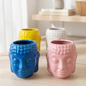 May include: A collection of ceramic Buddha head planters in various colors: blue, yellow, white, and pink. Each planter features a detailed face with closed eyes and a serene expression. These decorative planters are suitable for small plants or succulents.
