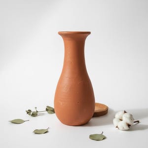 Puede incluir: Un jarrón de terracota con base redondeada y abertura acampanada. El jarrón es de un tono cálido y terroso y está rodeado de hojas secas, un pequeño posavasos de madera y una ramita de algodón. El jarrón mide unos 25 cm de altura.
