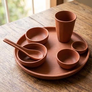 May include: A terracotta dinnerware set consisting of a large round tray, a tall drinking cup, three small bowls of varying sizes, and a long-handled spoon. Each piece features a matte, earthy reddish-brown finish and a simple, unadorned design.