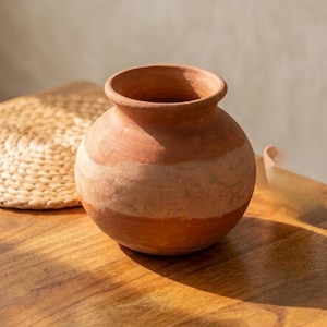 May include: A round terracotta pot with a wide opening and a short neck. The exterior features a natural reddish-brown clay colour with a distinct, lighter horizontal band encircling the middle section of the vessel, showcasing a textured surface.