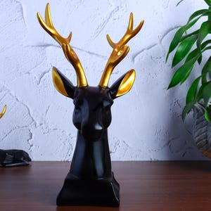 May include: A black deer head sculpture with gold antlers and ear detailing. The ornament is displayed on a wooden surface, with a plant in the background. The sculpture is a modern home decor item.