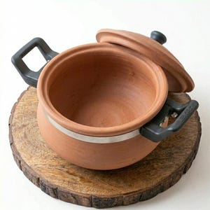 Pode incluir: Uma panela de terracota com acabamento em argila natural, apresentando duas alças laterais de plástico preto e uma tampa correspondente. Uma fina faixa metálica prateada circunda a parte superior externa da panela de borda circular.