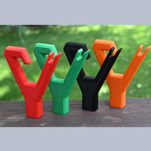 May include: Four colorful, Y-shaped tools in red, green, black, and orange, standing upright on a wooden surface. The tools have a unique design with a forked top. The background is a blurred view of green foliage.