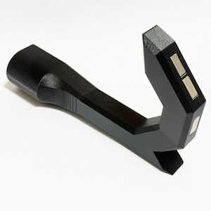 May include: A black, angular tool with a cylindrical handle and a right-angled head. The head features two small, rectangular silver inlays. The tool is made of a matte black material and sits on a white surface.